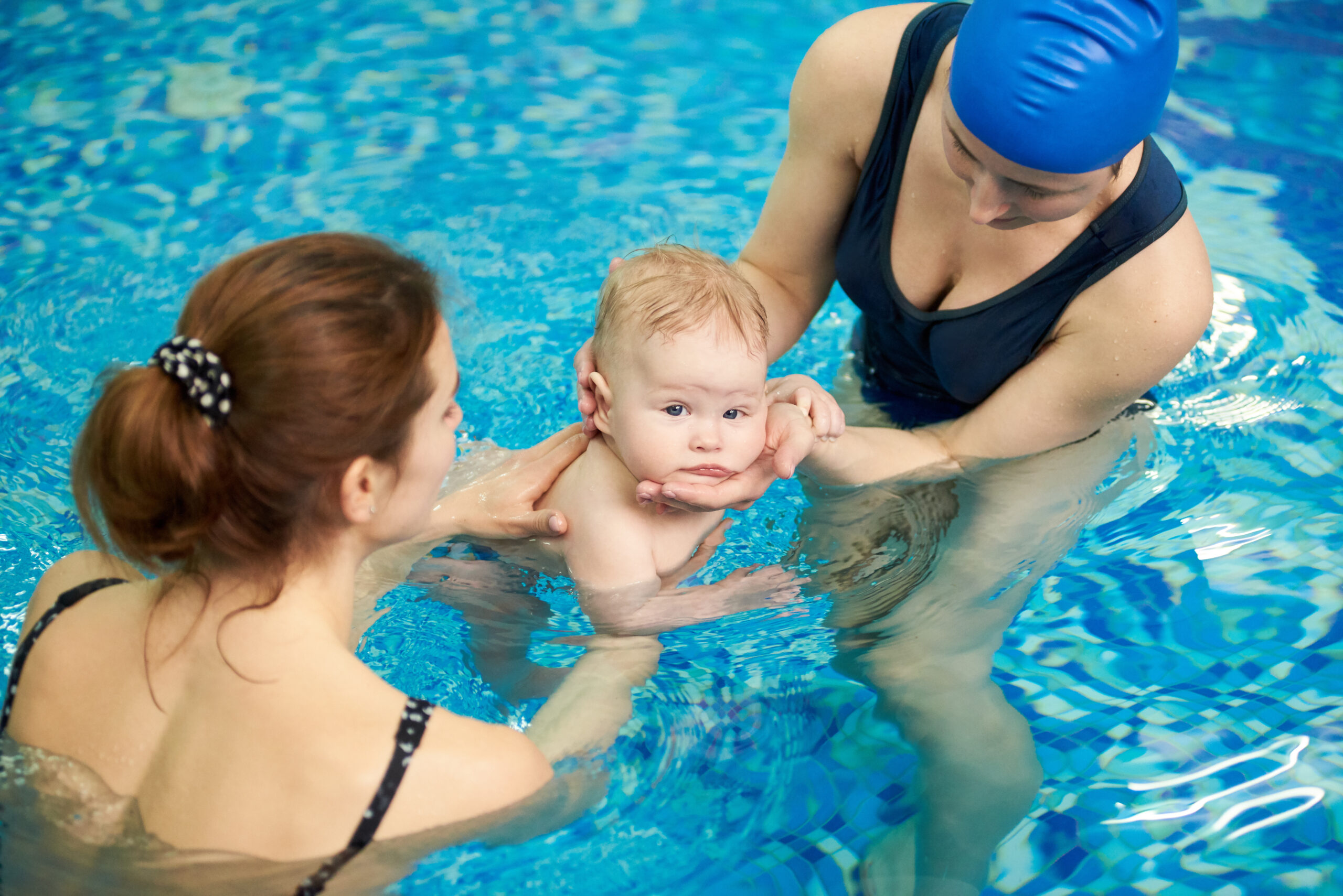 Swimmer little baby looking in camera while immersing in water at swimming classes in paddling pool. Young mother and female instructor teaching little child swimming and diving. Selective focus
