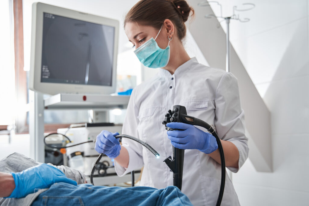 Female doctor wearing protective mask holding endoscope during gastroscopy in hand while little girl patient laying at the hospital bed. 