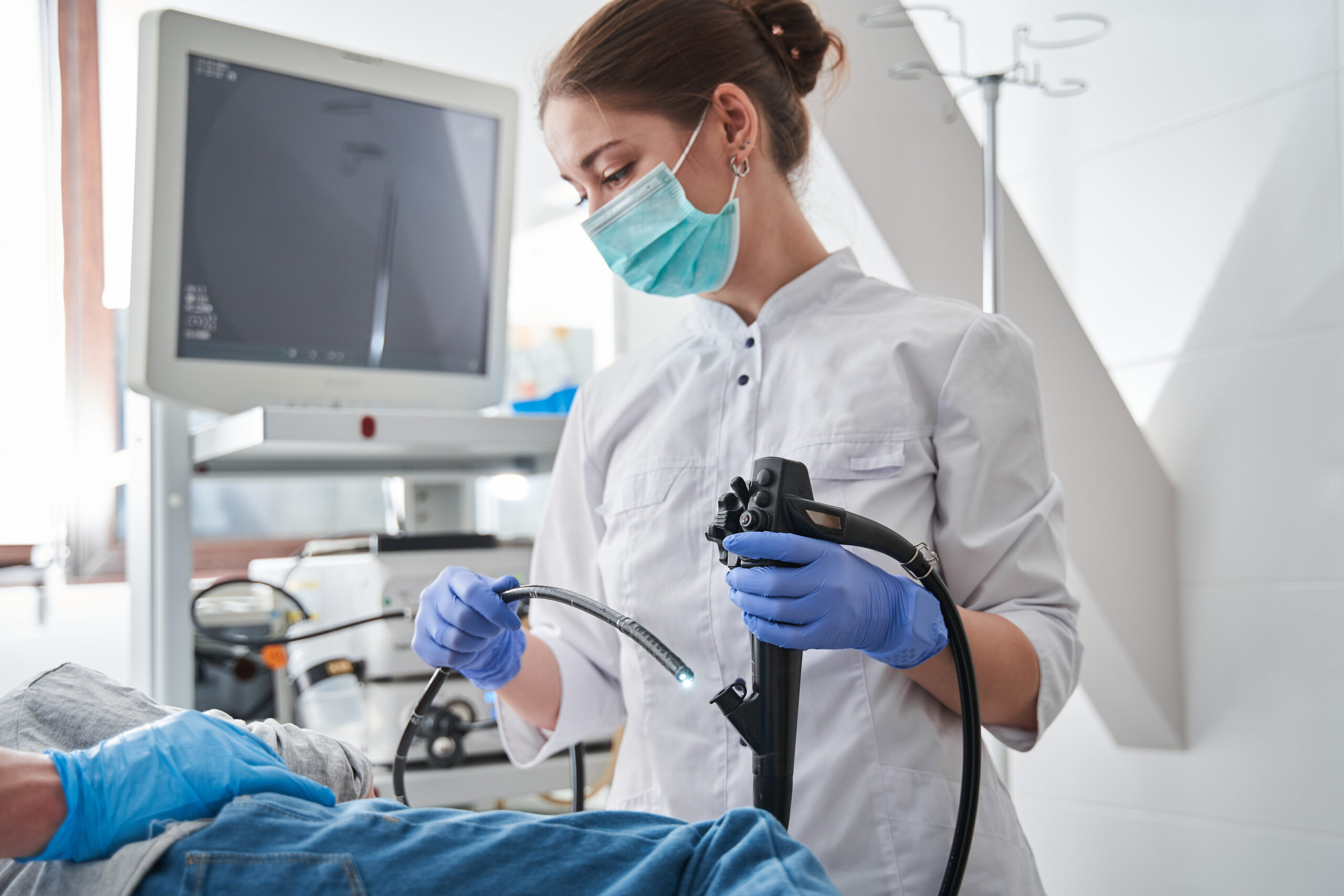 Female doctor wearing protective mask holding endoscope during gastroscopy in hand while little girl patient laying at the hospital bed.